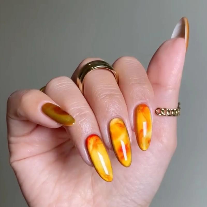 A hand with long almond-shaped amber tortoiseshell design nails wearing minimal gold jewelry - مدل ناخن مخملی