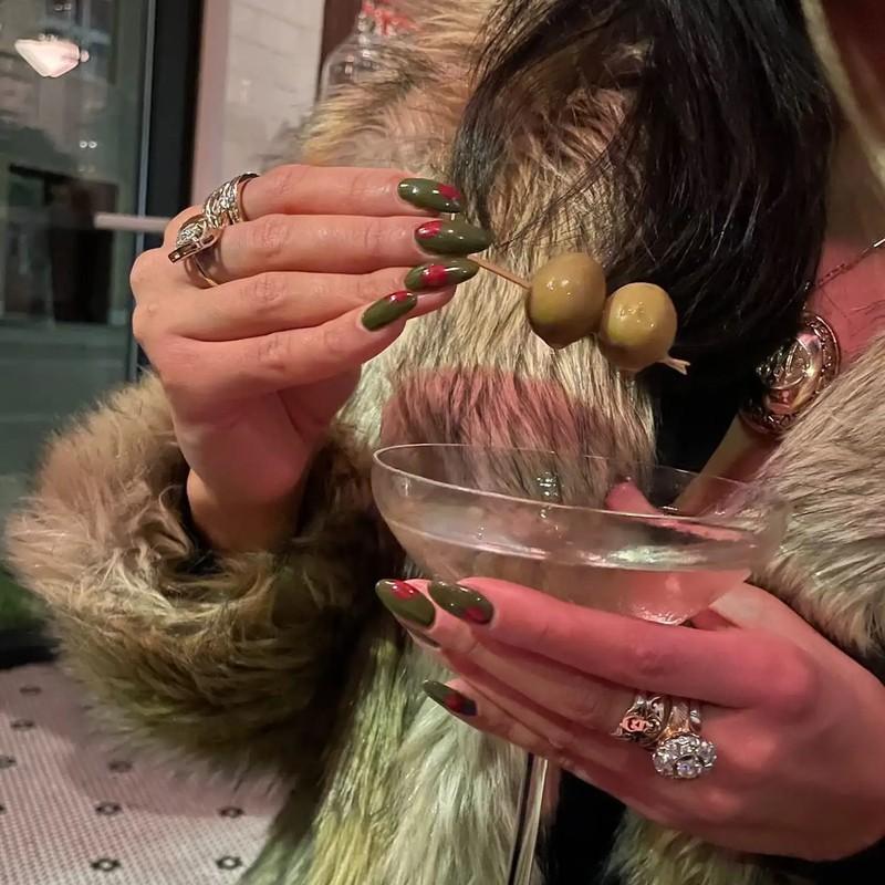 ایده ناخن زیتونی: A person holding two olives on a cocktail pick over a martini glass wearing ornate rings and a fur garment