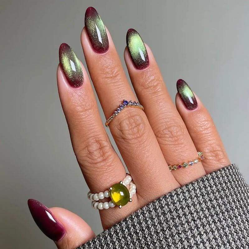 A hand with long ,shiny nails in a gradient wine red and green pattern adorned with multiple rings including a gem and pearl one - مدل ناخن مخملی