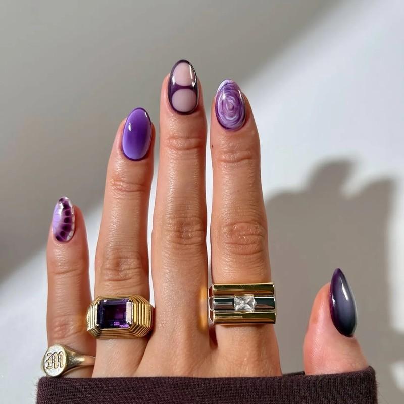Closeup of a hand wearing rings and having nails painted in various purple designs, مدل ناخن کوتاه پاییزی بنفش تک رنگ